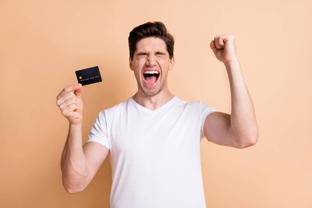 Photo of impressed guy closed eyes fist up open mouth shout yes hold debit card isolated on beige color background - Foto, Bild