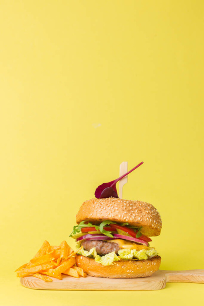 Sappige hamburger met kotelet, cheddar, arugula en gemengde saladebladeren. Serveer op een houten bord met aardappelchips. Close-up, kopie van de ruimte, verticaal, gele achtergrond - Foto, afbeelding