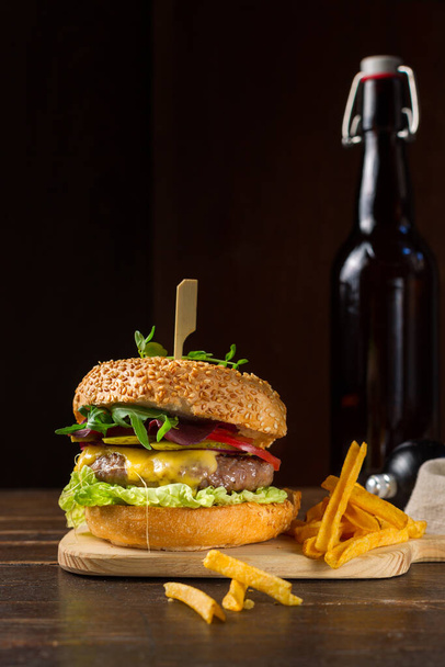 Köstlicher Burger mit Cheddar-Käse, Essiggurken, Tomaten und einer Mischung aus Salatblättern. Mit Chips und einer Flasche Bier servieren. Vertikal, zurückhaltend, rustikal. Ernährungskonzept - Foto, Bild
