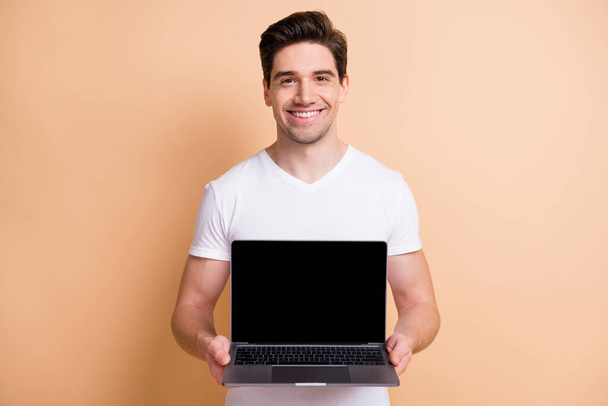 Portrait of nice young man toothy smile arms hold netbook showing display isolated on beige color background - Fotoğraf, Görsel