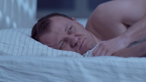 Acostado en la cama por la noche, un hombre no puede dormir, se cubre la cabeza con una almohada - Imágenes, Vídeo