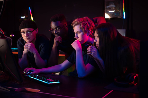 Zwei Jungs und ein Mädchen in medizinischen Masken in einem Computerclub unterstützen ihren Freund bei seinem Spiel - Foto, Bild