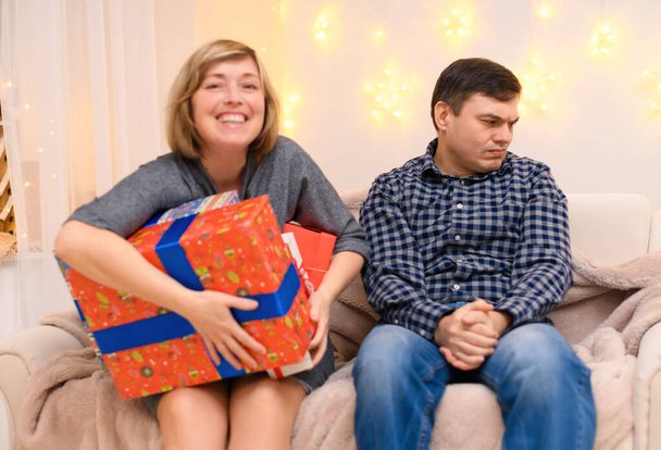 portrait of adult romantic couple, sitting with gifts on a couch at home, a woman takes away all the gifts from a man, the photo is like a joke, holiday lights on a wall - Photo, Image