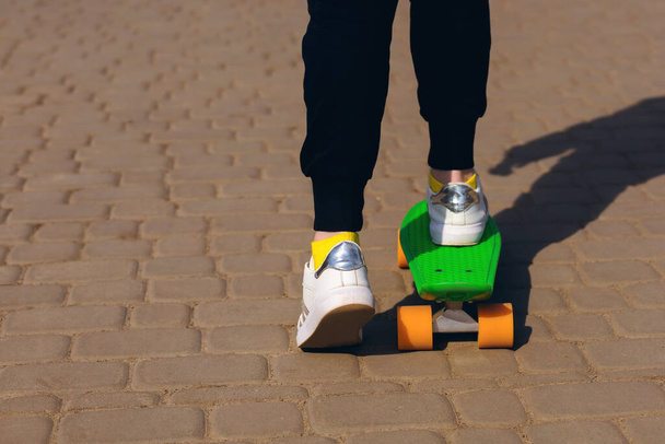 Ragazza adolescente cavalca uno skateboard o un penny board nel parco. Il bambino è vestito con abiti luminosi e sta imparando a cavalcare un penny board verde. Vista da vicino di gambe e skateboard - Foto, immagini