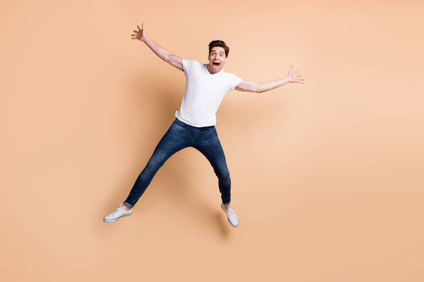 Full size photo of optimistic brunet man jump yell wear t-shirt jeans sneakers isolated on beige color background - Photo, image