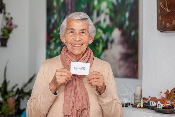Senior woman at her home holding her vaccination certificate after getting the Covid-19 vaccine in Colombia. Authentic vaccination certificate. Real patient - Фото, изображение