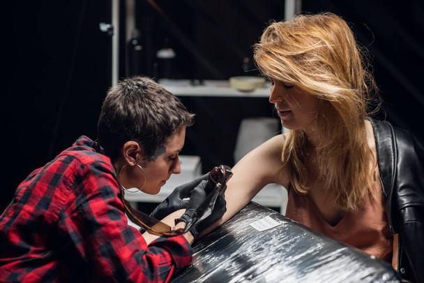 Moderno esperto maestro tatuaggio ragazza fa un tatuaggio sul braccio di una giovane ragazza preoccupata con i capelli rossi. - Foto, immagini
