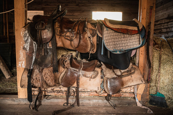 Ledersättel, weiche Decken, Stahlbügel und andere Reitutensilien im Holzstall auf der Ranch - Foto, Bild