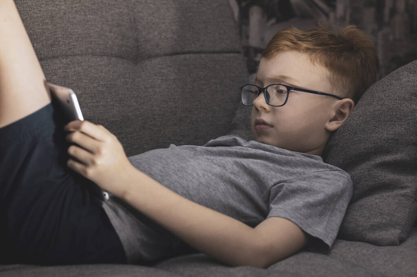 A boy with glasses for vision lies on the couch and looks into the tablet. - Fotografie, Obrázek