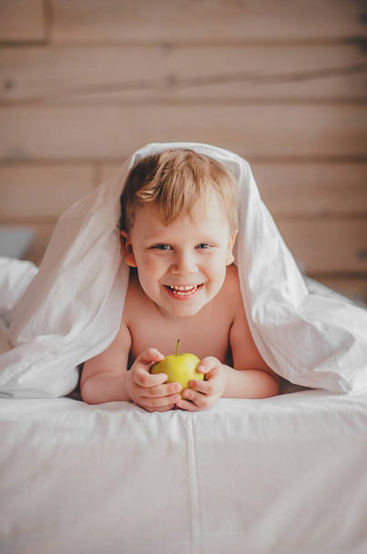 Een 5-jarige blanke jongen ligt onder de deken, houdt een gele appel in zijn handen en glimlacht vrolijk. Kijkt in de camera. - Foto, afbeelding