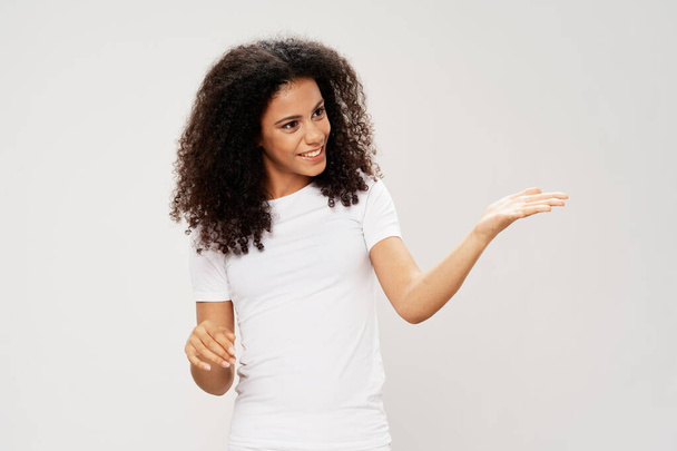 curly-haired woman of African appearance in a white T-shirt gestures with her hands on a light background Copy Space - Foto, Imagem