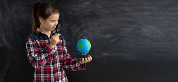 young caucasian girl schoolgirl or student stands near the chalk blackboard. He is holding a globe with the help of a magnifying glass, looking for something on the world map. Baner - Φωτογραφία, εικόνα