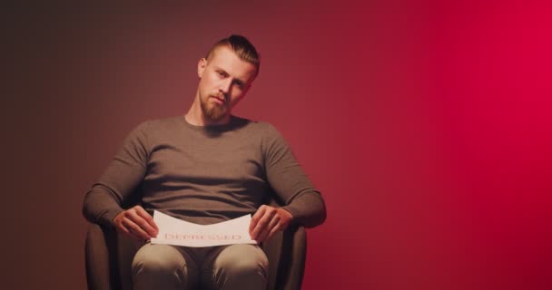 Stressaantunut nuori mies, jolla on paperilevy istuu nojatuolissa tummaa taustaa vasten - Materiaali, video