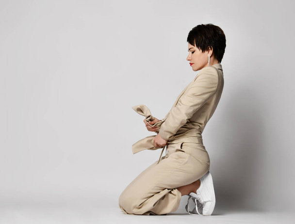 Adult woman in beige business casual pantsuit and sneakers stands on knees holding belt. Stylish business female wear - Photo, image