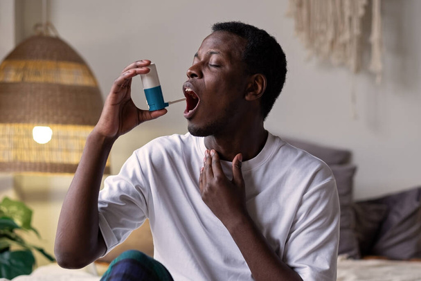 Young african man using asthma inhaler sitting at home - Фото, изображение