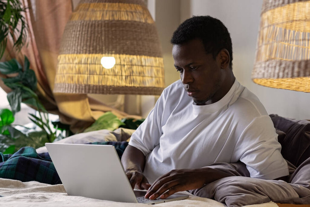 Молодой серьезный африканский человек, печатающий дома на ноутбуке - Фото, изображение