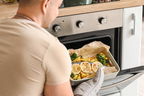 Young man taking out of oven baking dish with fish and potatoes in kitchen - Photo, Image