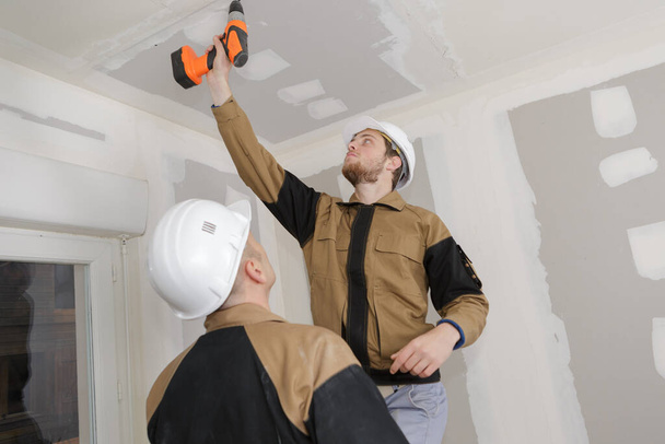 young handsome builder with a drill sets plasterboard ceiling - Φωτογραφία, εικόνα