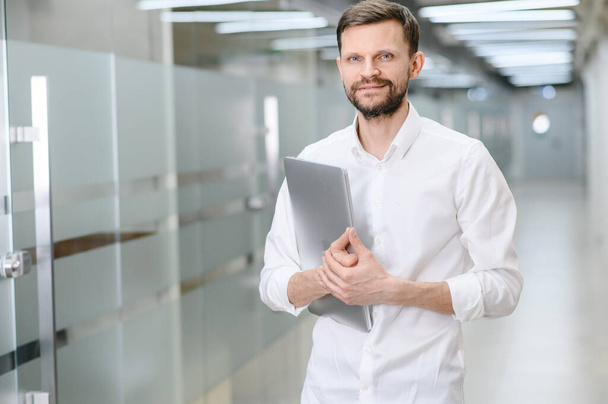 portrait of a handsome young man holding a laptop and smiling happily at the camera, copy space. A man in the background of the office - Foto, Imagen
