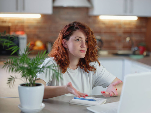 Μια όμορφη κοκκινομάλλα κάθεται στην κουζίνα στο τραπέζι με το λάπτοπ της. μελέτη σε απευθείας σύνδεση με skype δάσκαλος, ευτυχισμένη νεαρή γυναίκα μαθαίνουν γλώσσα, μακρινή εκπαίδευση - Φωτογραφία, εικόνα