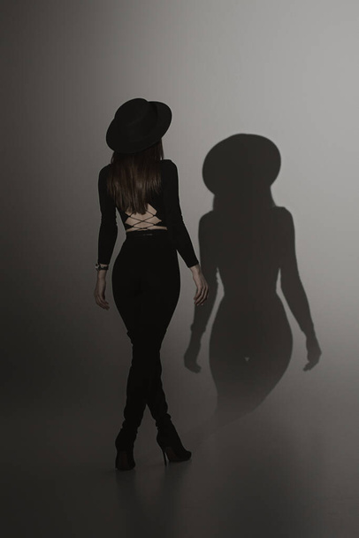 Καυτή κομψή γυναίκα στο στούντιο. Νεαρή γυναίκα με μαύρη στολή και καπέλο. Το κορίτσι στέκεται με την πλάτη της - Φωτογραφία, εικόνα