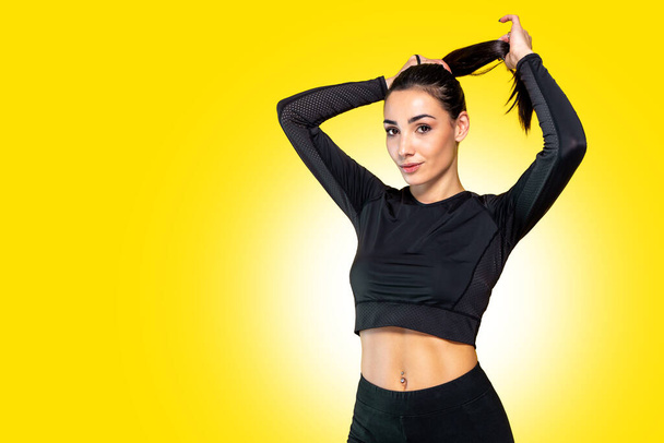 Beautiful active young sportive caucasian athlete woman training in black sportswear outfit with yellow background behind - Studio shooting with pretty brunette posing relaxing after sport training - Photo, Image