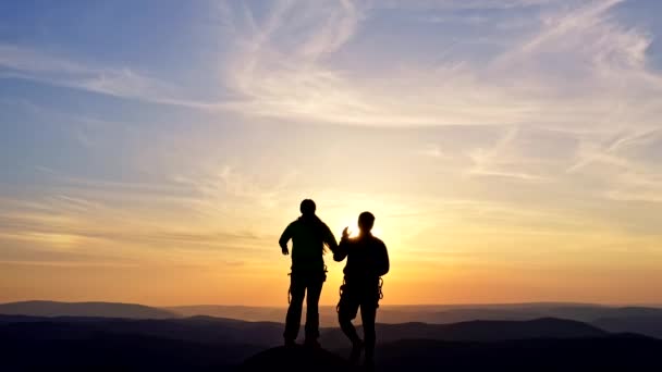 Um jovem casal alegremente levanta as mãos enquanto está em pé no topo de uma montanha ao pôr do sol. - Filmagem, Vídeo