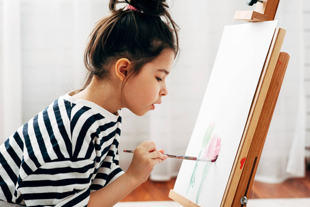Widok z boku portret skoncentrowanej dziewczynki malującej pędzlem na papierze na sztaludze w domu. Urocze dziecko siedzące na podłodze i rysujące w jej pokoju podczas zamknięcia. - Zdjęcie, obraz