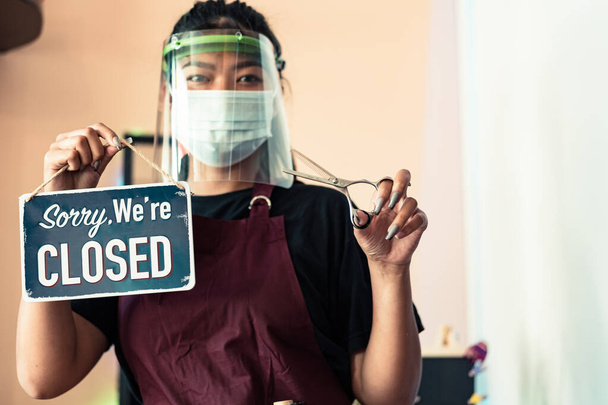 Aranyos ázsiai női fodrász szemfedővel, védő maszk fekete pólóban és kötény egy fém lemez SORRY vagyunk zárva, hogy lezárja intézkedés. A koronavírus fogalma. - Fotó, kép