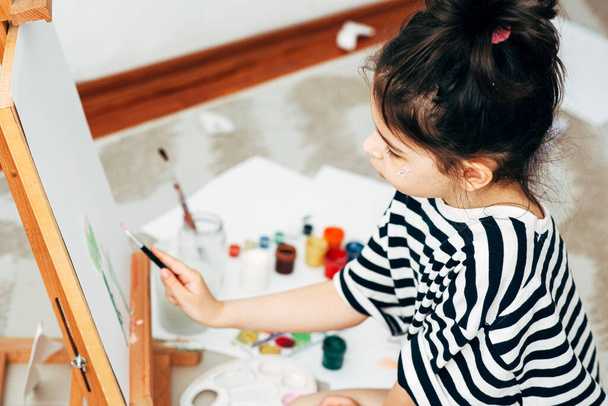 Overhead image of a cute little girl kid painting on the paper on an easel at home. A happy kid sitting on the carpet and drawing in her room during quarantine. - Foto, Imagem