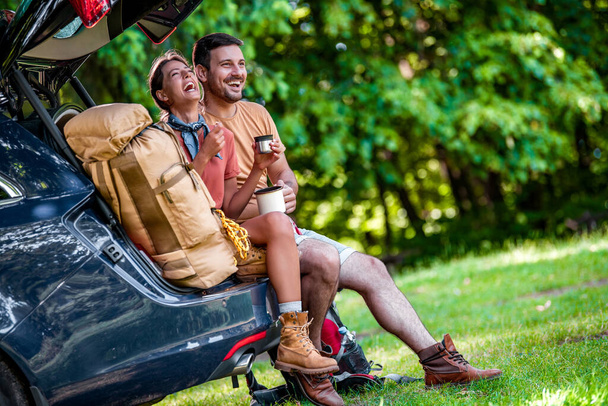 Пара туристов сидят в багажнике и пьют кофе или чай. Летом. Путешествия, отдых и люди. - Фото, изображение