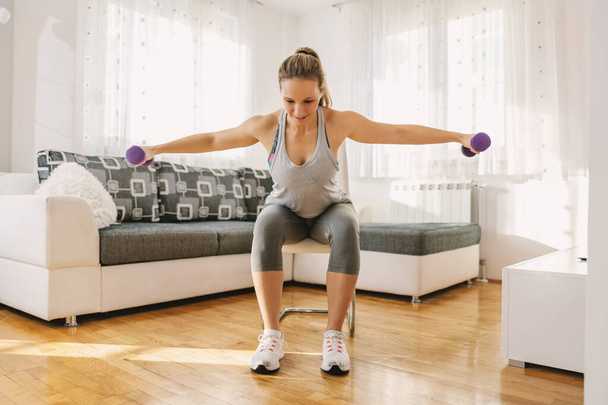 Smiling muscular woman in shape sitting on the chair at home and doing fitness exercises for arms with dumbbells. - Photo, Image