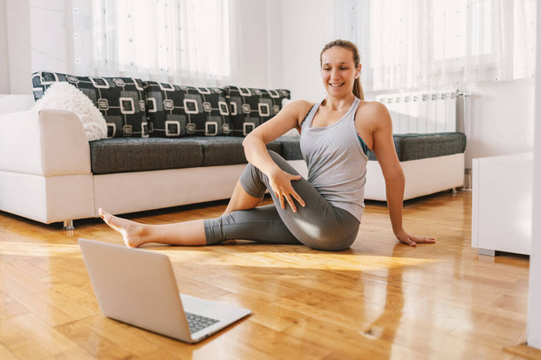 Glimlachende yoga instructeur zittend op de vloer thuis in Lord of the Fishes yoga pose en het hebben van online klasse over laptop. - Foto, afbeelding