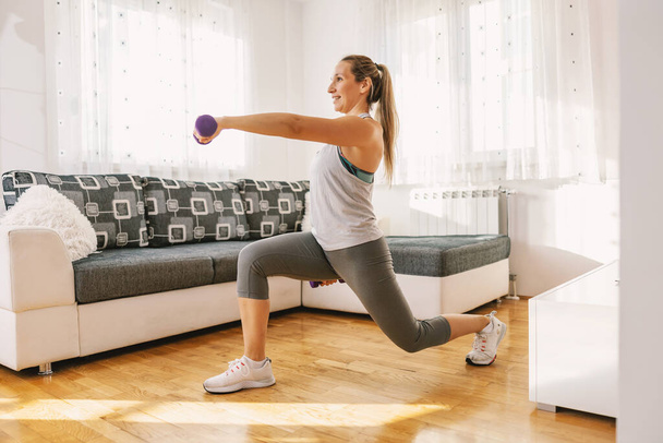 Smiling muscular sportswoman in shape doing lunges while holding dumbbells. - Fotó, kép
