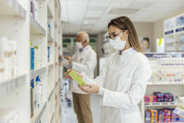 A female pharmacist standing by a drug shelf at the time of the corona virus. She is wearing a face mask and a white uniform. She holds a package of medicines in his hands and reads the declaration - Foto, Imagem