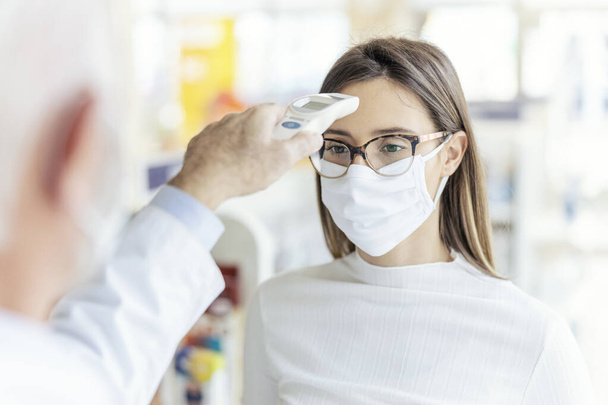 Messung der Körpertemperatur mit einem digitalen Thermometer, Schutz vor Coronaviren. Eine Nahaufnahme des Oberkörpers einer Frau mit Brille und Schutzmaske im Gesicht. Gesundheitscheck - Foto, Bild