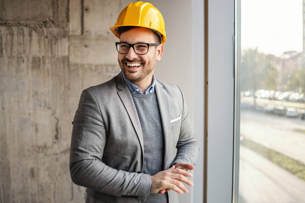 Usmívající se architekt v obleku stojící vedle okna ve stavebnictví a dívající se jinam. - Fotografie, Obrázek