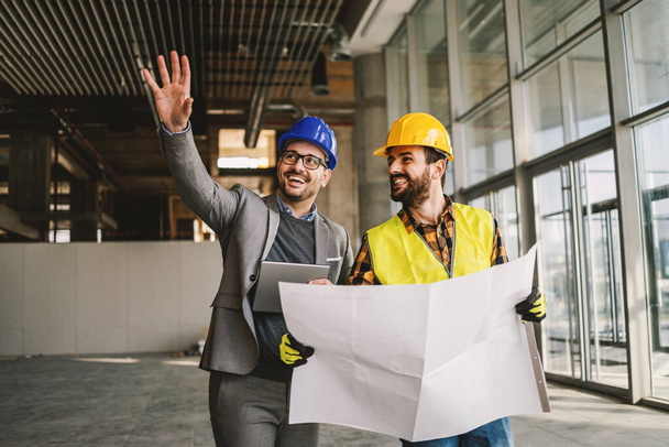 Smiling architect holding tablet and showing to construction worker something he imagined. Construction worker holding blueprints. Construction site interior. - Photo, Image