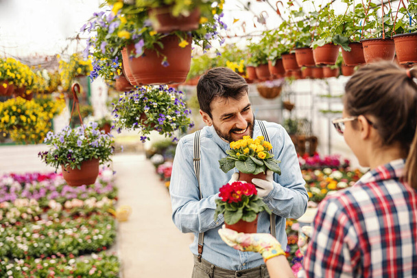 Dos emprendedores sonrientes enamorados coqueteando y sosteniendo macetas con flores. A su alrededor hay todo tipo de flores de colores. Interior del invernadero. - Foto, Imagen