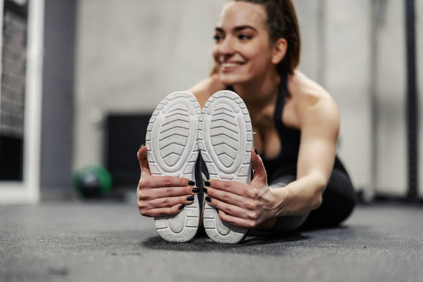 Muscle stretching in spot training. A female person sits on the gym floor and stretches her leg and foot muscles. Women's hands hold the sole of the sneaker and stretch the leg muscles, worming up - Фото, изображение