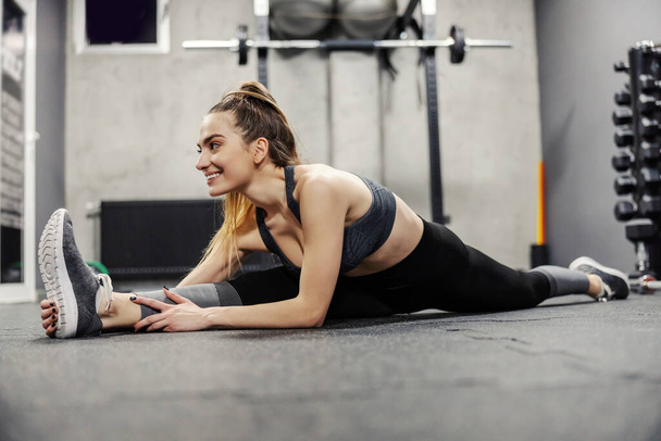 Muscle flexibility and hip mobility. Fit woman in sportswear and good body shape stretches legs muscles on the floor of an isolated gym. Enjoyment and smile in training, ponytail - Foto, Imagem
