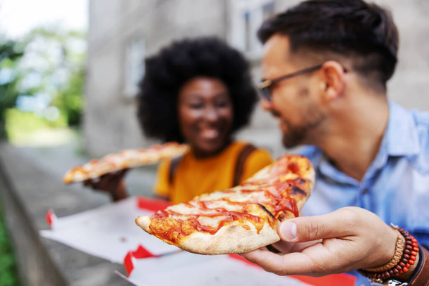 Closeup of young multicultural hipster couple sitting outdoors and eating pizza. Selective focus on man'd hand. - Photo, Image