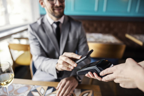 Online payment of restaurant bills. A man in a business suit with a tie sits at a table in a restaurant and leans his phone on the terminal to pay for a meal during lunch time. Online e-banking - Foto, imagen