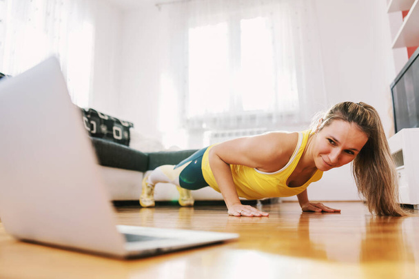 Fitness εκπαιδευτής κάνει pushups στο σπίτι και έχοντας online τάξη με τους μαθητές της. - Φωτογραφία, εικόνα