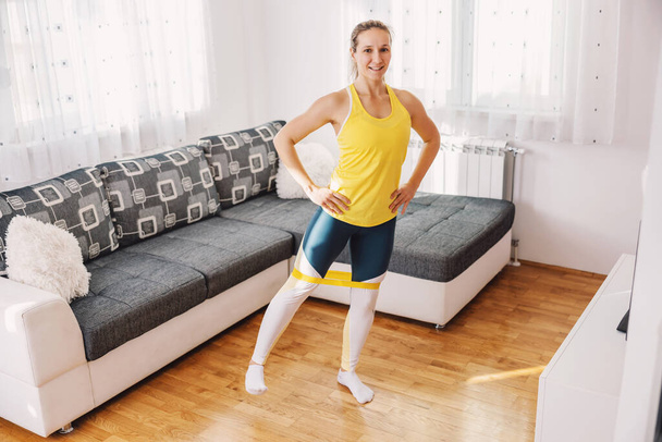 Smiling fit sportswoman standing at home with hands on hips and stretching power rubber with legs. She is doing fitness exercises. - Photo, Image