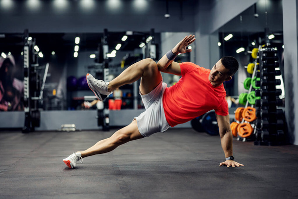 A motivated male sportsperson does a side plank for core and ads exercises with one hand and one leg up in the gym fitness studio. Well-being, gym challenge - Foto, Imagem