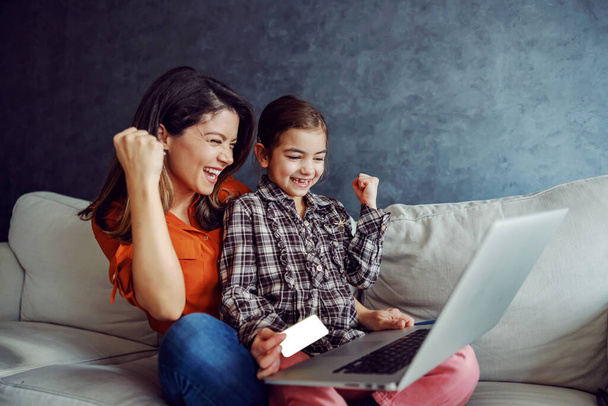 Uśmiechnięta matka i córka siedzą na kanapie i używają laptopa do zakupów online. Córka trzyma kartę kredytową. Mieli udany zakup, więc są rozradowani i trzymają pięści. - Zdjęcie, obraz