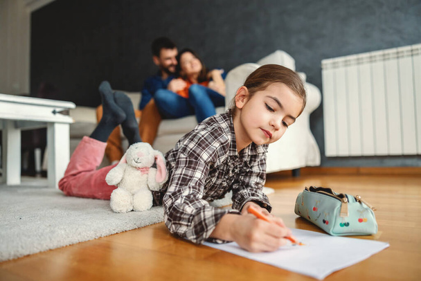 Child lying down on stomach on the floor drawing. In blurry background parents hugging. Happy family. - Fotoğraf, Görsel