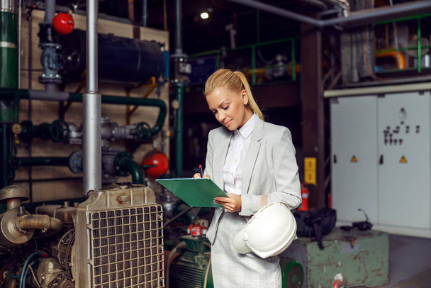 Inspection in the heating plant. A blonde businesswoman stands in a heating plant, holding a clipboard and checking machines. She is dressed in a business suit and holds a protective helmet in hand - Photo, Image