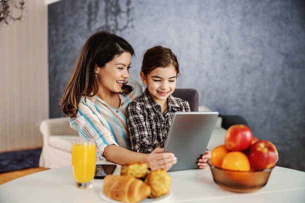 Smiling mother and daughter sitting at dining table in the morning and using tablet. Happy family watching cartoons on tablet. - Foto, Imagem